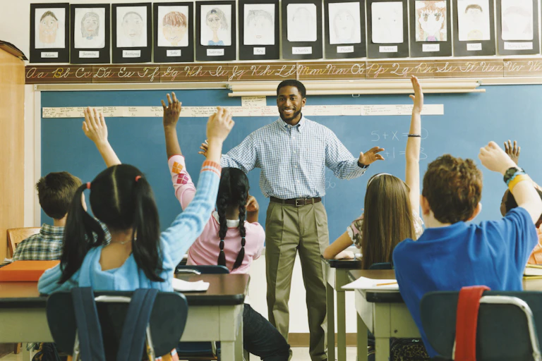 a black male adult stands at the front of a classroom as school-age children sitting at desks raise their hands
