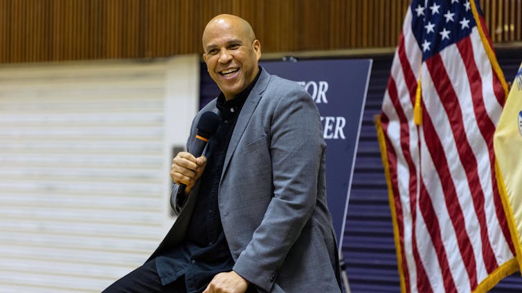 Trump, interrumpido: discurso de 25 horas del Cory Booker de 25 horas para la acción colectiva 2 El hombre sostiene el micrófono y las sonrisas. Detrás de él hay una bandera americana