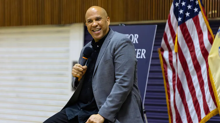 A man holds a microphone and smiles. behind him is the US flag