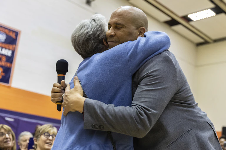 A man holding a microphone embraces a grey-haired woman.