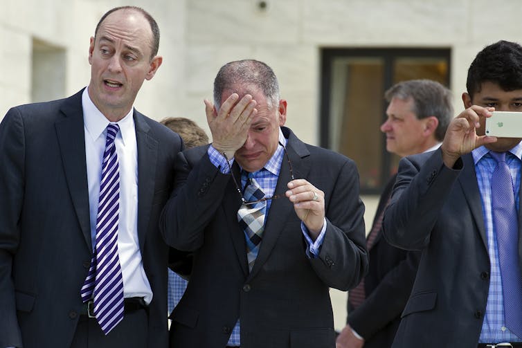 A man in a suit wipes tears from his eyes as he stands next to another man in a suit in a hallway.