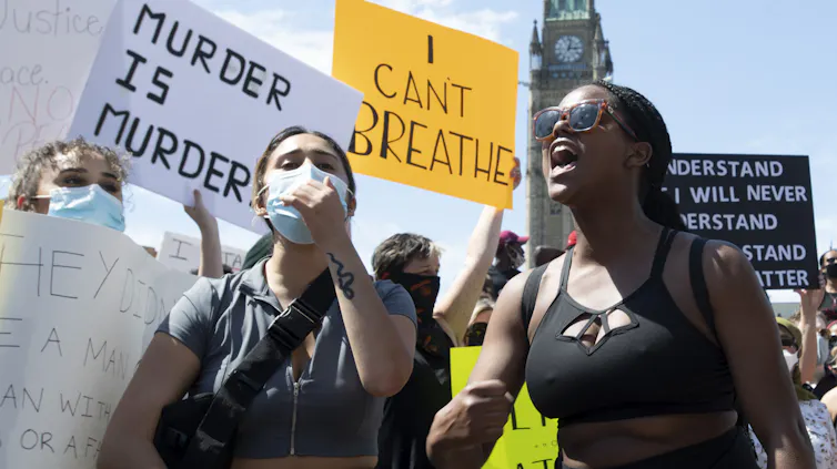 two women stand at a protest in front of signs. one says murder is murder the other says I can't breath