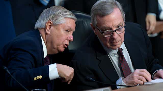 Two older men in business suits lean toward each other.