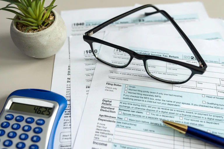 An individual income tax form, a calculator, a pair of glasses, a pen and a small houseplant are arrayed on a table.