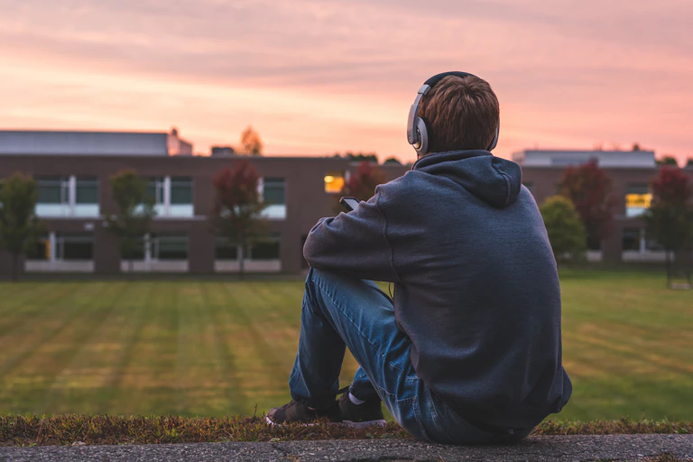 Teen boy listening to headphones sits on grass hill looking at sunset.
