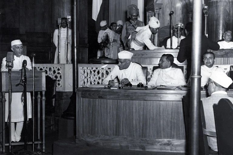 Jawaharlal Nehru, the first prime minister of India, speaks at a podium in the Indian parliament in Delhi, wearing a traditional Indian white coat and hat. Other parliamentarians listen to his speech.