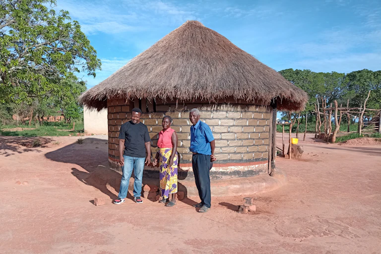 Two men and a woman standing outside a one roomed round thatched house