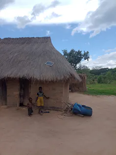 A small house on a dusty piece of land with a tiny solar panel on the thatched roof, only enough to charge a phone. Two small children stand in front.