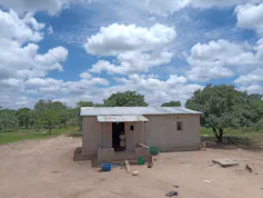 A house made of concrete blocks stands alone in the middle of a remote and dusty area with trees in the background