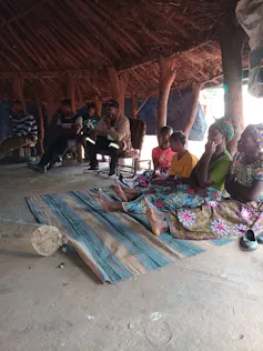 A group of people sit on a mat and plastic chairs under a thatched roof, discussing solar power