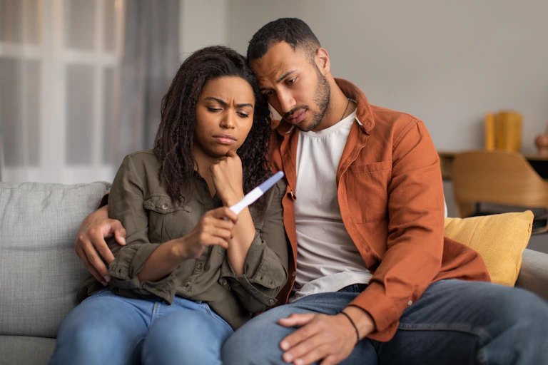 A man and a woman on a sofa, looking at a pregnancy test and looking disappointed.