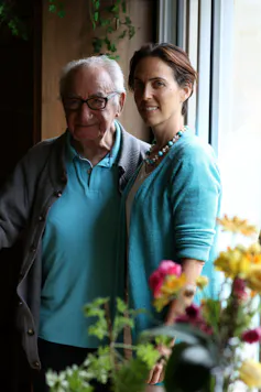 A man with white hair, glasses and a turquois polo shirt stands with a woman with short brown hair and a turquoise sweater.