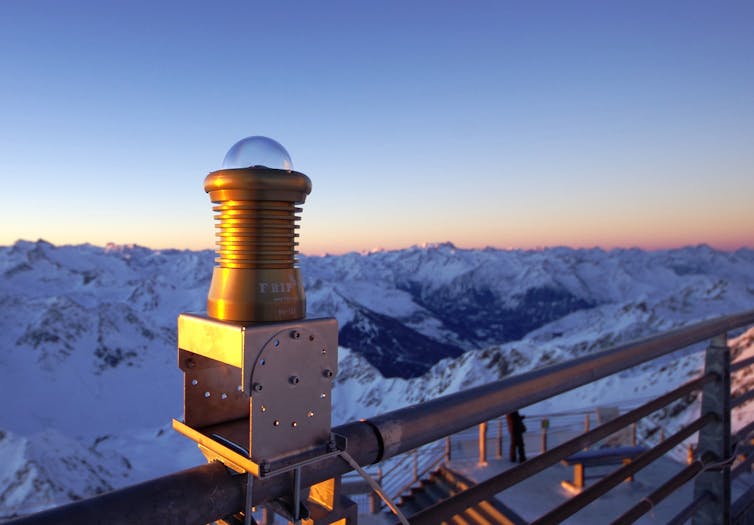 A camera, which looks like a glass sphere, attached to a metal stand on a railing overlooking mountains.