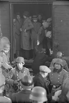 A black and white photo of men in coats and hats crammed into a cargo car, as soldiers in hard hats stand outside.