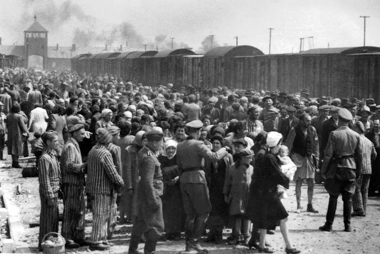 A black and white photograph of a crowd of people and soldiers outside with smoke, gates and train cars in the background.