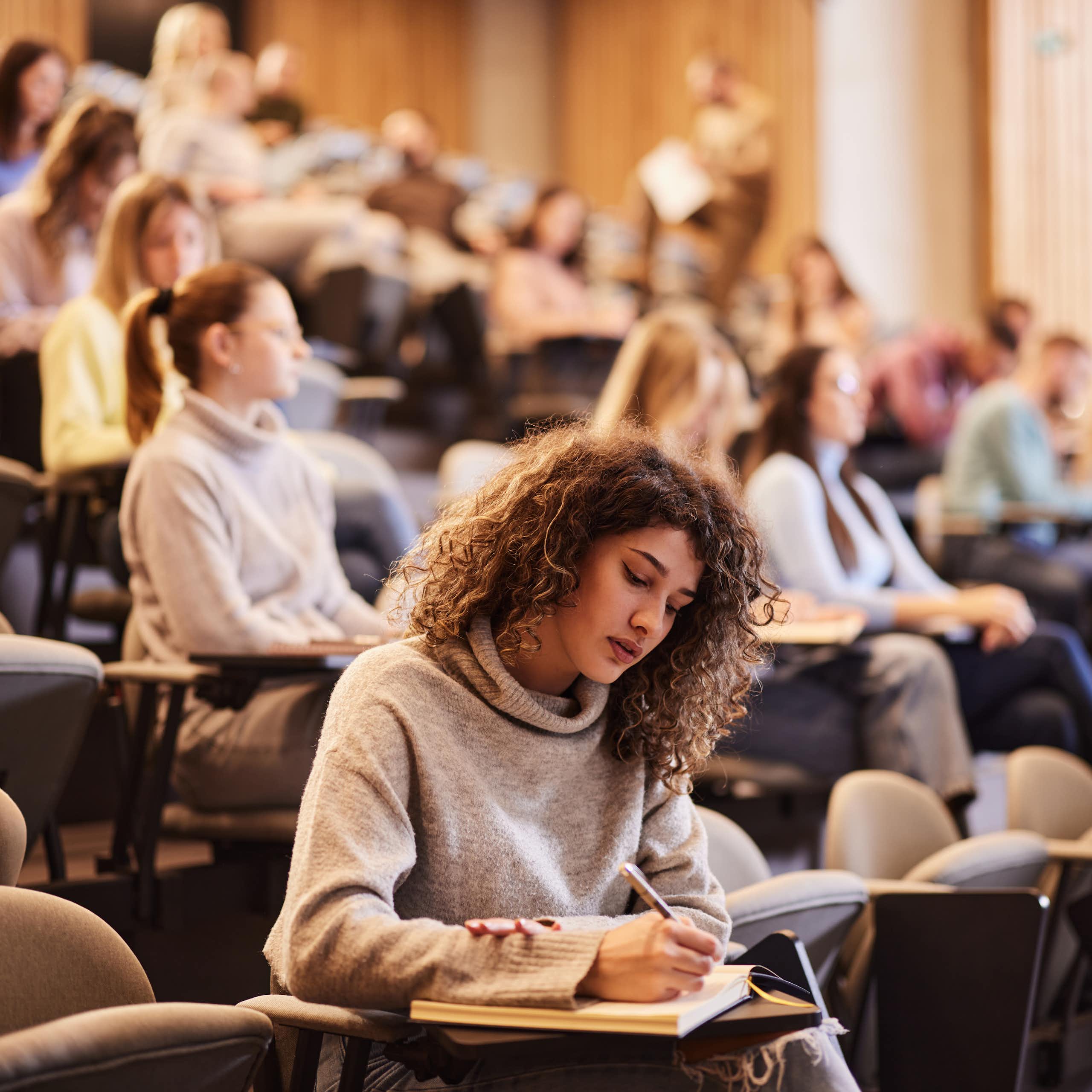 Female college student writing an exam during a class at lecture hall. Her classmate are in the background.