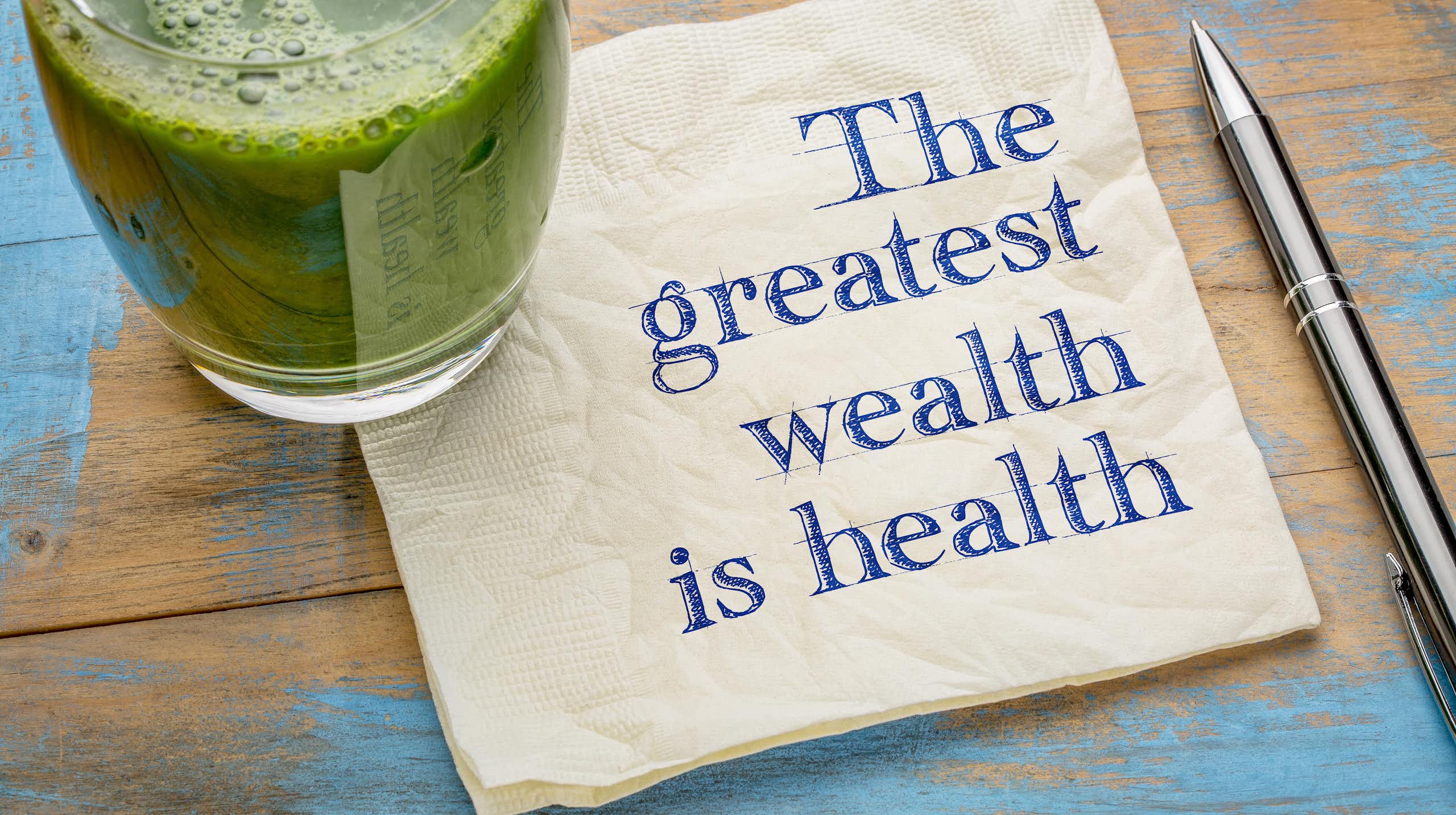 A glass of green juice sitting on a napkin that reads 'The greatest wealth is health.'