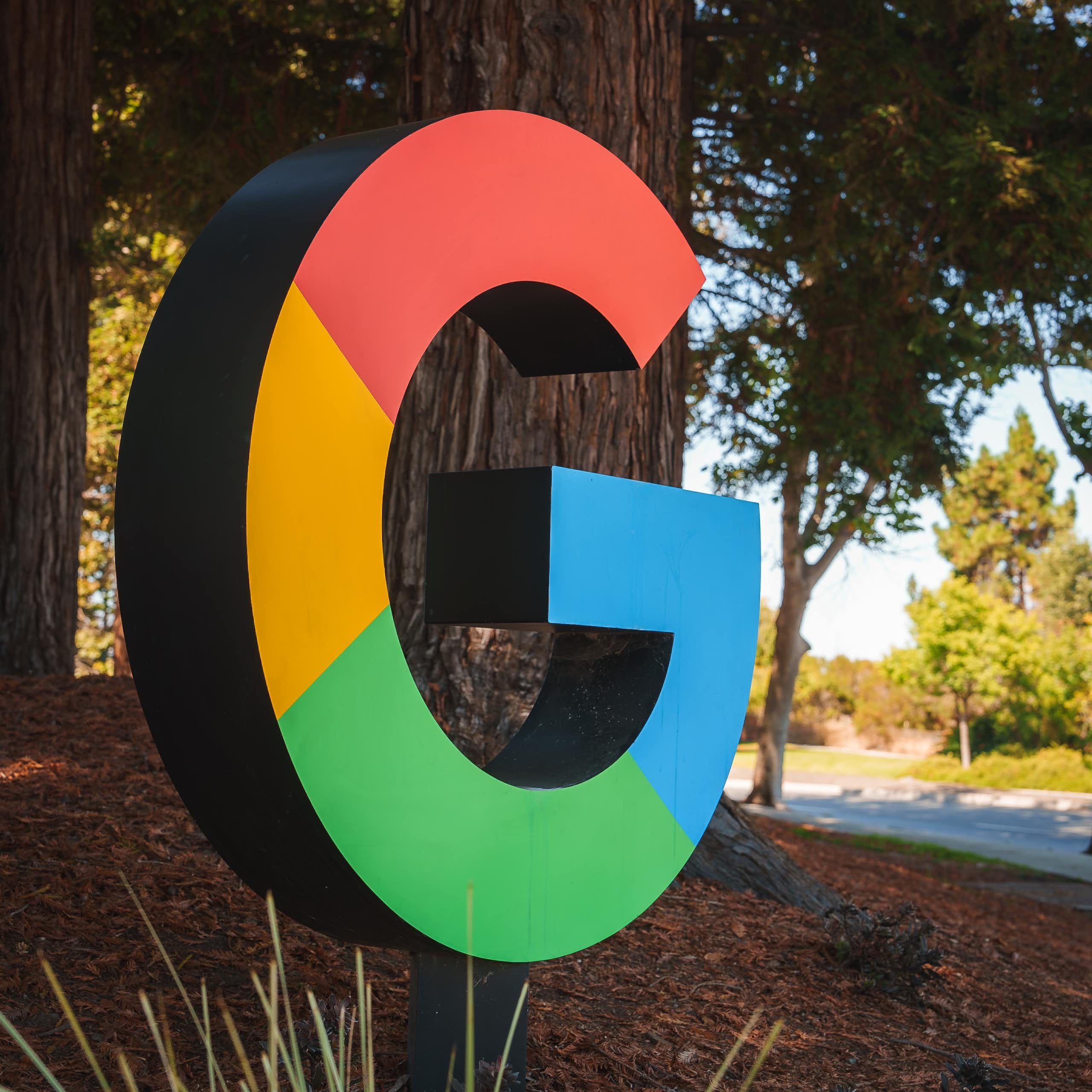 A large, multicoloured capital letter G appears near a road in a wooded setting.