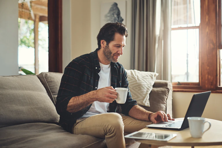 Man using laptop while sitting on a sofa holding a mug.