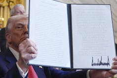 Um homem com cabelo e pele brancos tingidos de laranja segura um documento em frente ao seu rosto.