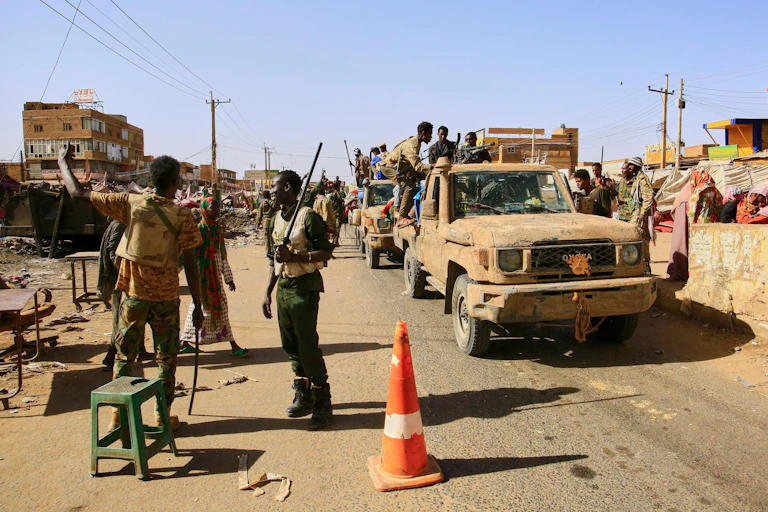 Armed troops from the Sudan Armed Forces in a convoy of pick-up trucks on a road beside which there is a small make-shift market.
