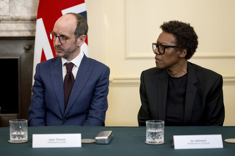 Dos personas se sientan en una mesa en la discusión con la bandera británica detrás de ellos.