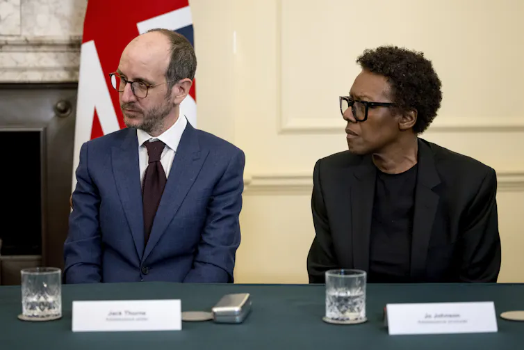 Two people seated at a table in discussion with the British flag behind them.
