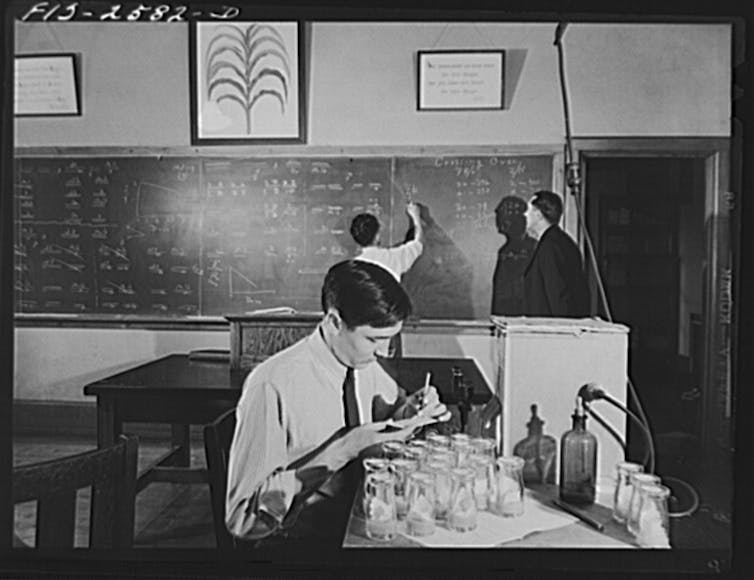 seated man in shirt and tie makes notes above jars, two other men at chalkboard with a poster of corn above