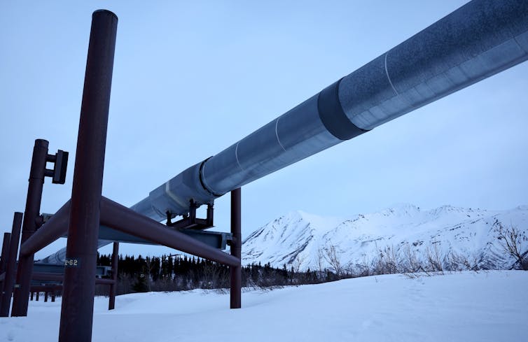 A large metal pipe sits on a metal base, with snow-covered mountains in the background.
