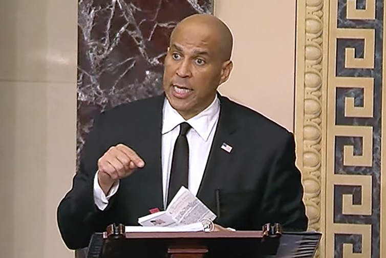 A Black man speaks on stage in front of a podium.