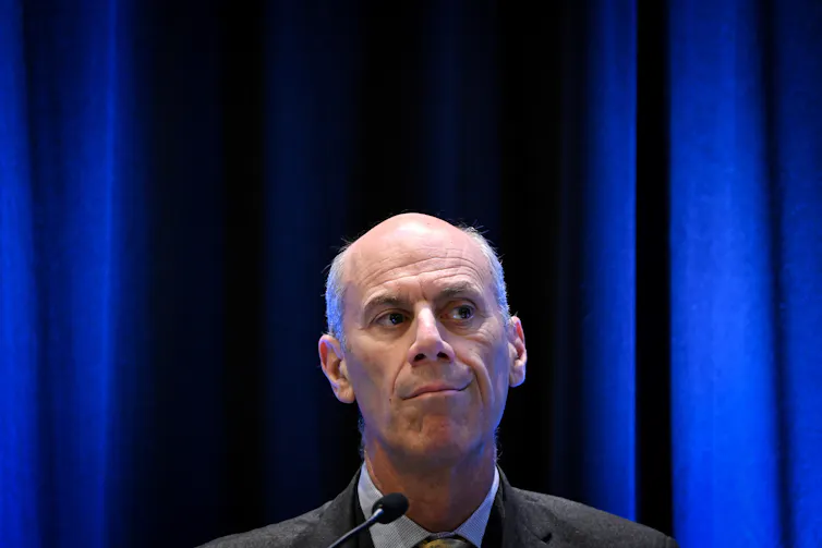A balding, serious looking man sitting in front of blue curtains.