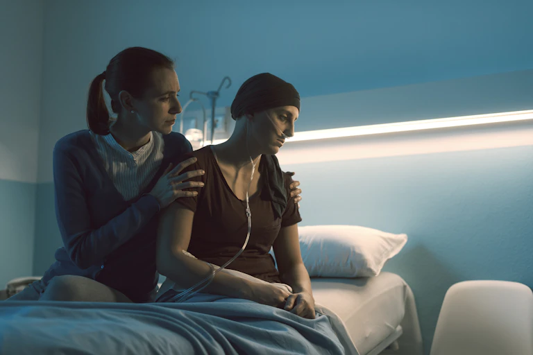 Despondent cancer patient wearing knit hat and nasal cannula sitting on hospital bed with another person holding their shoulders