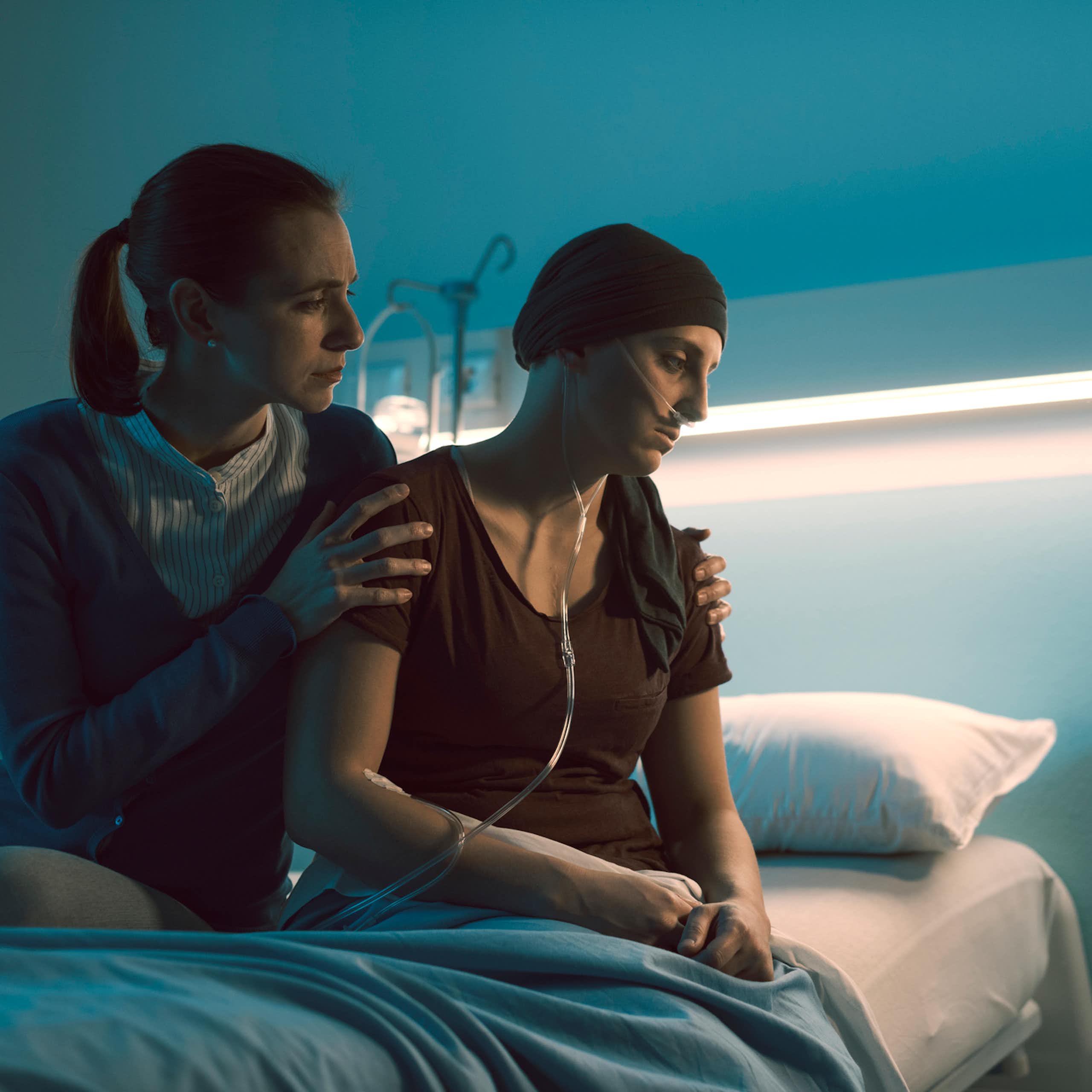 Despondent cancer patient wearing knit hat and nasal cannula sitting on hospital bed with another person holding their shoulders