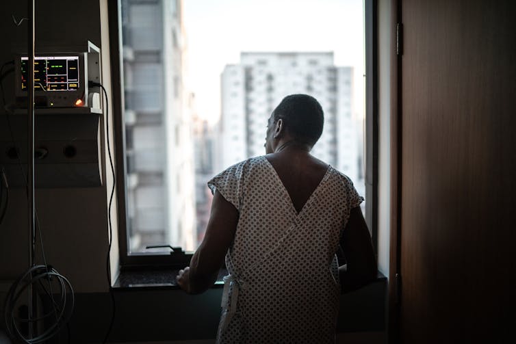 Patient in hospital gown looking out window, hands lightly braced against the sill