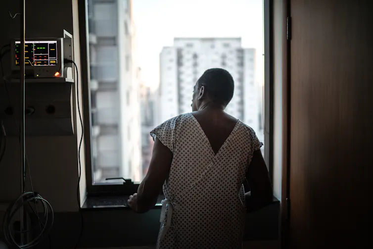 Patient in hospital gown looking out window, hands lightly braced against the sill