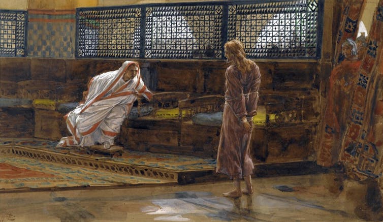 A man in a togalike robe leans forward on a bench as he talks to a thin man standing before him with bound arms.