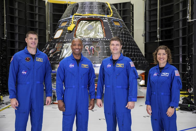 Four astronauts in blue NASA jumpsuits standing in front of a conical metal capsule.