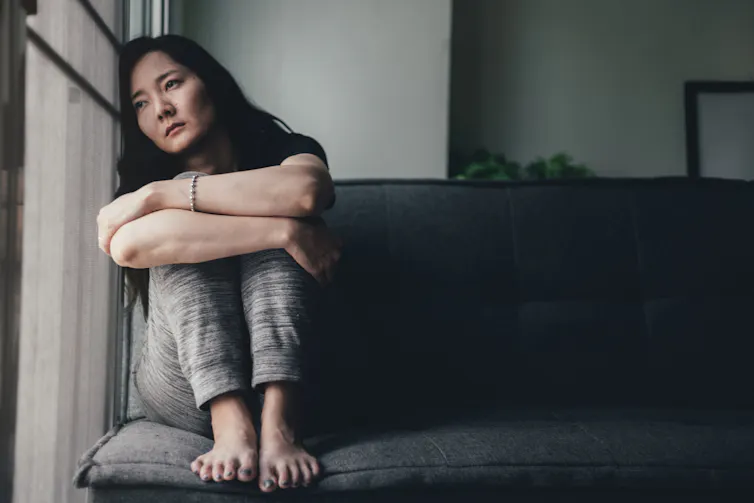A young woman sits leaning against a window with her arms wrapped around her knees