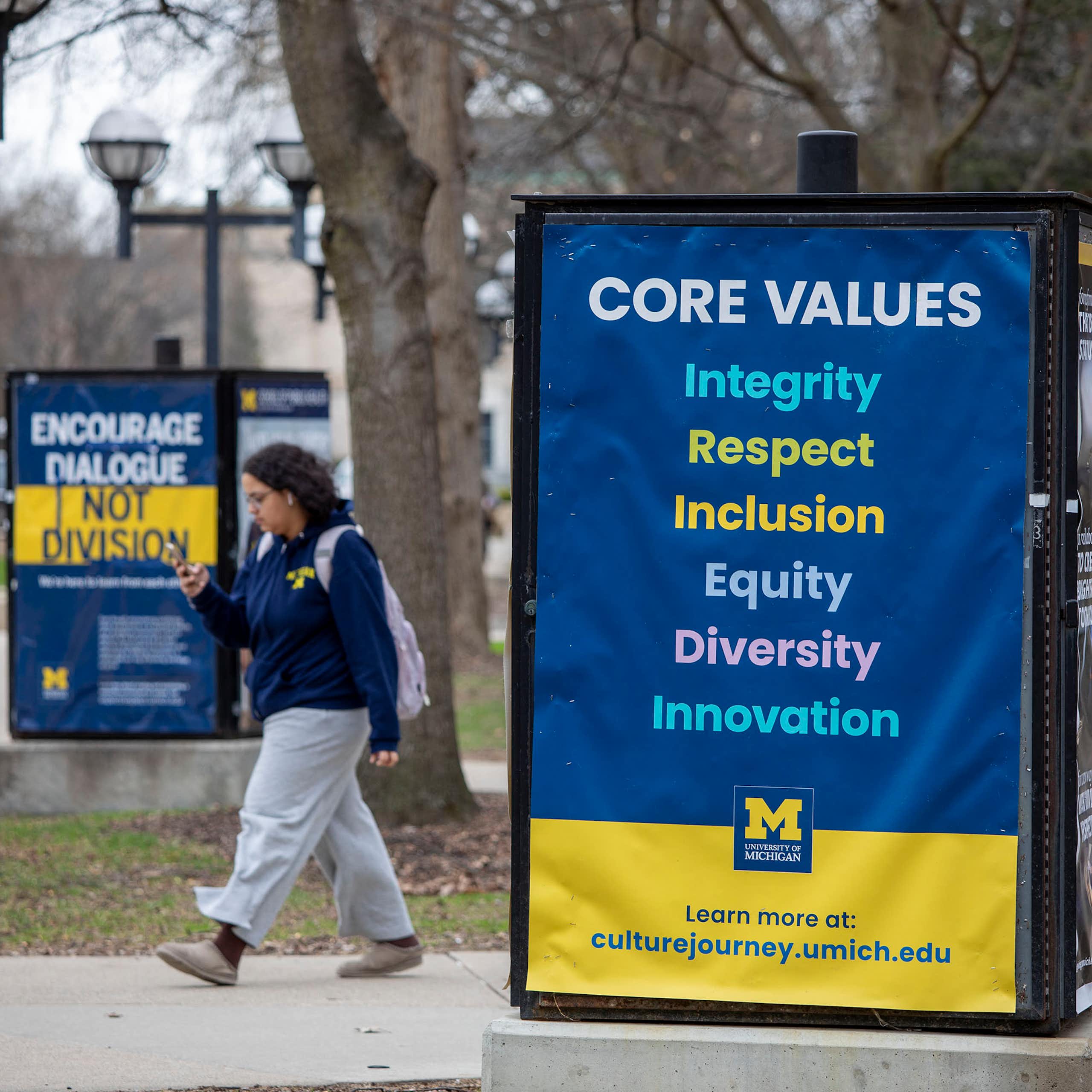 A person wearing a blue top walks on a cement path next to an advertisement for the core values of the University of Michigan.