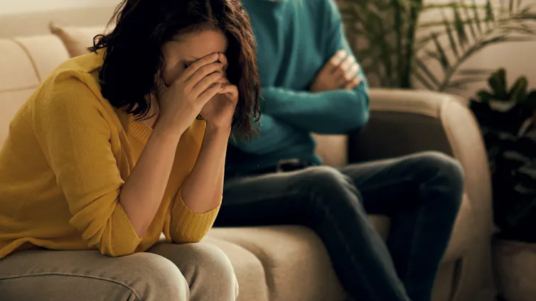 A woman sits on a couch with her head in her hands while another person, only seen from the neck down, sits beside her with their arms crossed