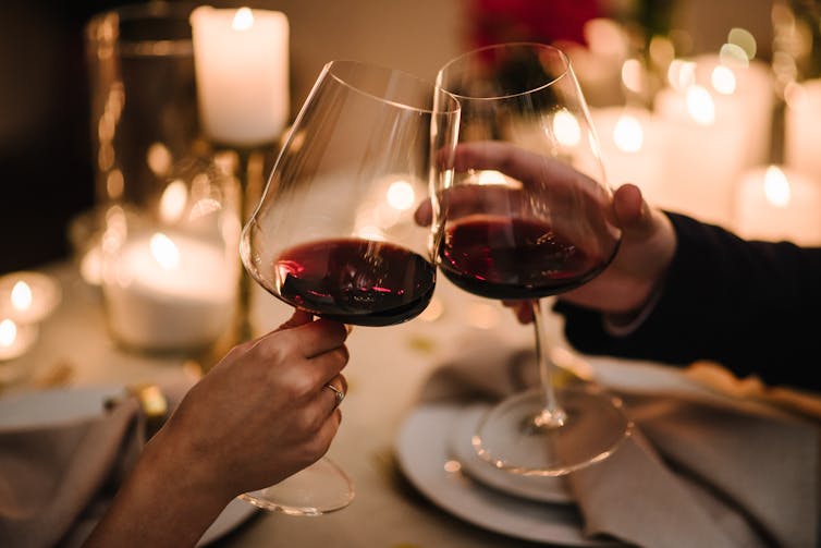 Two people knock their glasses of red wine together over a candlelit table