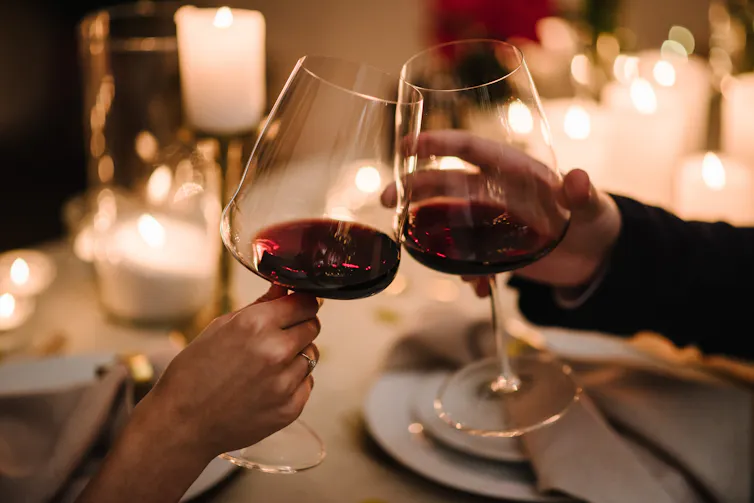 Two people knock their glasses of red wine together over a candlelit table