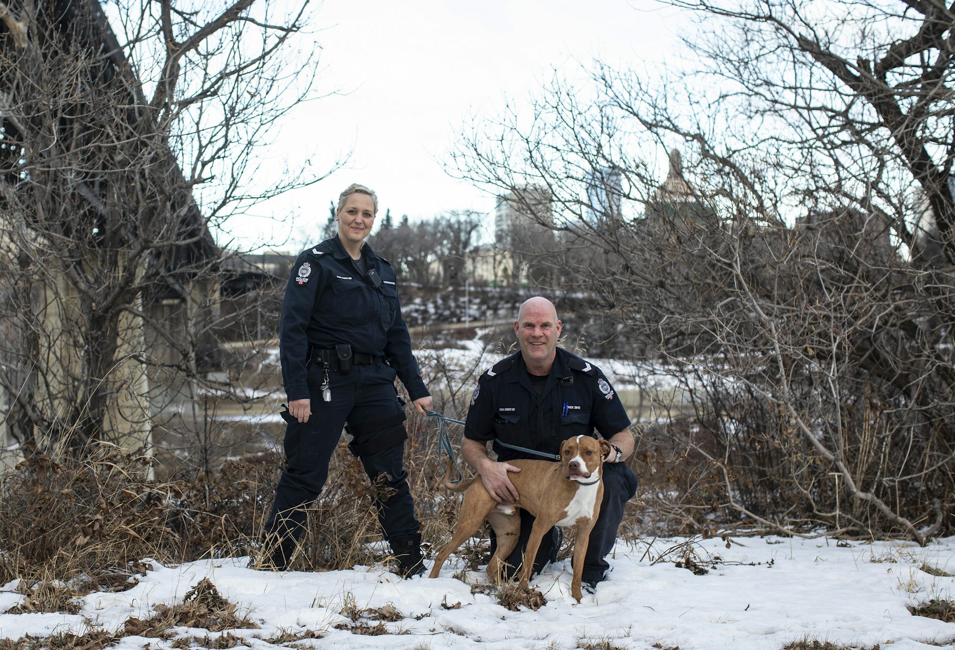 Outside by trees and in snow, a woman stands and a man squats, both in police uniform, next to a brown and white dog