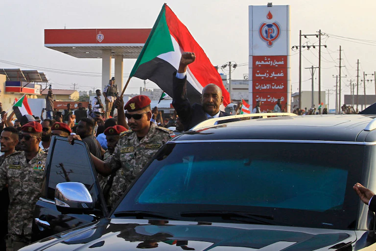 A man waving from the door of a vehicle as a crowd surrounds him near a petrol station with a red, white, black and green flag waving in the middle of the crowd