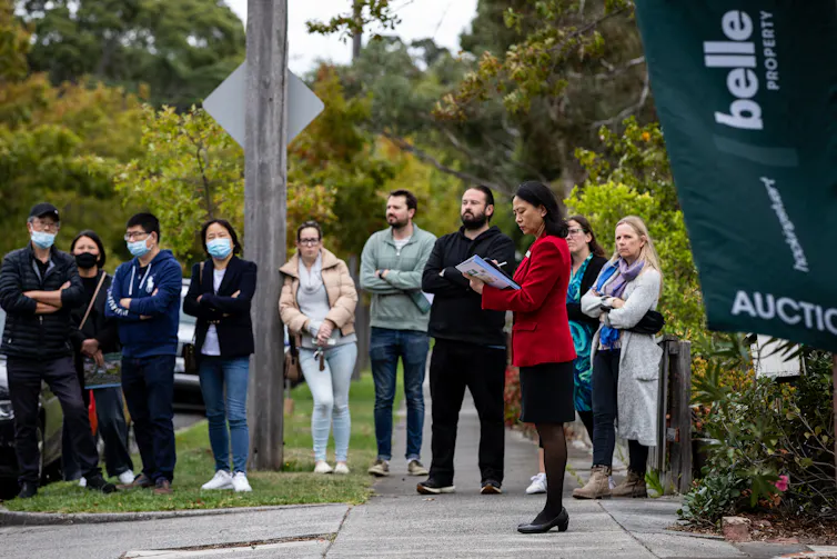 Several people standing on a footpath in front of a house auction banner and an female auctioneer wearing a red jacket and black skirt
