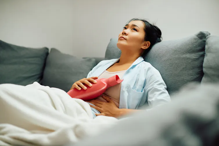 A woman holding a hot water bottle to her stomach lying on a couch.