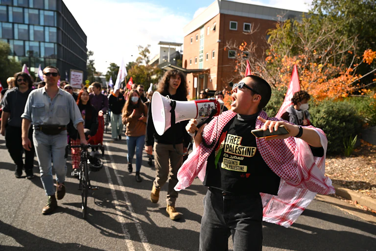 People walk down a street. A man shouts in a megaphone.