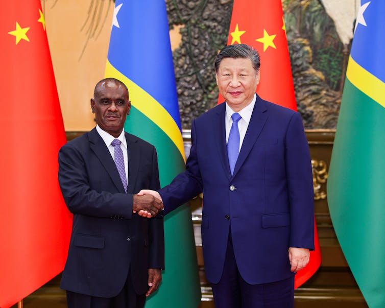 two men in suits shake hands in front of flags