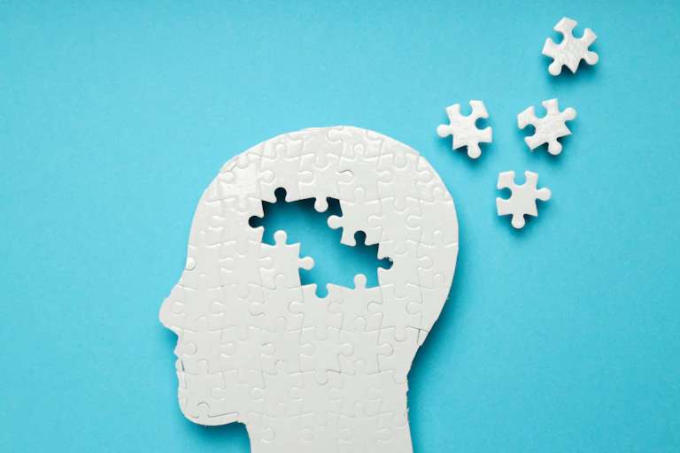 A head in profile made of white puzzle pieces. In the centre of the head, puzzle pieces are missing and have been scattered next to it.