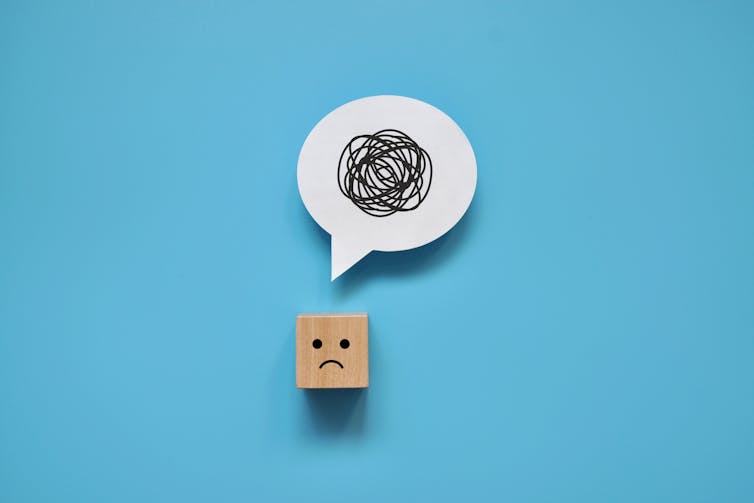 A wooden cube with a frowny face. It has a speech bubble above it that has black scribbles.